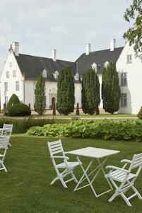 A stately white mansion with dark roofs is surrounded by neatly trimmed green hedges and tall trees. In the foreground, a manicured lawn features several white folding chairs and a table, creating an elegant and peaceful outdoor setting.