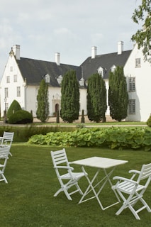A stately white mansion with dark roofs is surrounded by neatly trimmed green hedges and tall trees. In the foreground, a manicured lawn features several white folding chairs and a table, creating an elegant and peaceful outdoor setting.