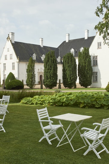 A stately white mansion with dark roofs is surrounded by neatly trimmed green hedges and tall trees. In the foreground, a manicured lawn features several white folding chairs and a table, creating an elegant and peaceful outdoor setting.