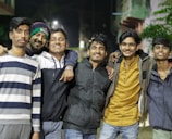 A group of six young men stands closely together, smiling widely, in a nighttime outdoor setting. They are dressed in casual winter clothing, with jackets and sweaters, and appear to be friends enjoying each other's company.