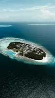 An aerial view of a small, lush island surrounded by turquoise waters. The island contains a community of buildings, bordered by white sandy beaches and a protective reef circling the clear ocean.