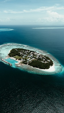 An aerial view of a small, lush island surrounded by turquoise waters. The island contains a community of buildings, bordered by white sandy beaches and a protective reef circling the clear ocean.