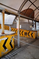 A weathered, enclosed structure featuring several yellow barriers with black arrow markings. The barriers are rusted, and there are various signs in a non-English language. The area appears to be an old or abandoned checkpoint or toll booth with metal frames and a faded roof covering.