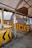 A weathered, enclosed structure featuring several yellow barriers with black arrow markings. The barriers are rusted, and there are various signs in a non-English language. The area appears to be an old or abandoned checkpoint or toll booth with metal frames and a faded roof covering.