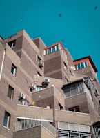 A series of modern, angular apartment buildings with brick facades and several balconies. The structures have a layered, stair-step design with air conditioning units attached to the walls. The sky is clear and turquoise, and a few birds are flying in the background.