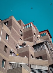 A series of modern, angular apartment buildings with brick facades and several balconies. The structures have a layered, stair-step design with air conditioning units attached to the walls. The sky is clear and turquoise, and a few birds are flying in the background.