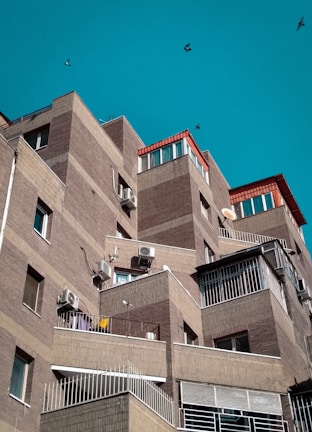 A series of modern, angular apartment buildings with brick facades and several balconies. The structures have a layered, stair-step design with air conditioning units attached to the walls. The sky is clear and turquoise, and a few birds are flying in the background.