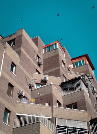 A series of modern, angular apartment buildings with brick facades and several balconies. The structures have a layered, stair-step design with air conditioning units attached to the walls. The sky is clear and turquoise, and a few birds are flying in the background.