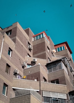 A series of modern, angular apartment buildings with brick facades and several balconies. The structures have a layered, stair-step design with air conditioning units attached to the walls. The sky is clear and turquoise, and a few birds are flying in the background.