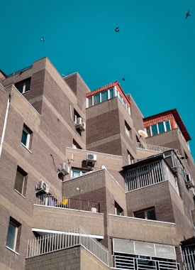 A series of modern, angular apartment buildings with brick facades and several balconies. The structures have a layered, stair-step design with air conditioning units attached to the walls. The sky is clear and turquoise, and a few birds are flying in the background.