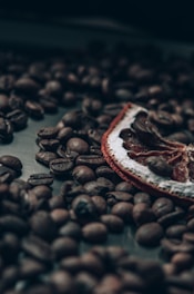 A close-up view of roasted coffee beans scattered on a surface with a dried citrus slice partially visible among the beans. The image has a dark and moody aesthetic with a soft focus effect, emphasizing the textures and shapes of the beans and citrus peel.