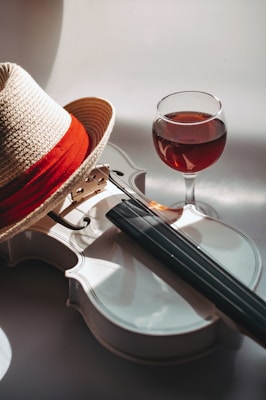 A white violin with a wooden bridge is placed on a light surface, accompanied by a straw hat with a red band and a glass of red wine. Soft shadows and reflections give the setting a warm and inviting feel.