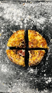A round baked pastry topped with sesame seeds, cut into four equal quarters. It rests on a dark surface dusted with flour, creating a rustic baking ambiance.