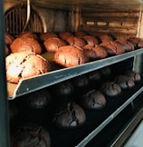 Rows of freshly baked, round chocolate cookies are cooling on metal racks inside an oven. The cookies have a cracked surface and a rich, dark brown color, indicating a deep, chocolate flavor. The oven's interior has a warm, inviting glow.