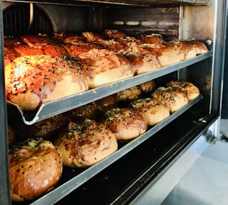 Several trays of freshly baked bread rolls are in an oven. The rolls are golden brown with a glossy finish, and some are topped with sesame or poppy seeds.