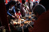 A group of people, including adults and children, are gathered around a traditional water purification fountain. They are holding wooden ladles used to scoop and pour water as part of a cleansing ritual. The atmosphere appears busy and communal, with everyone actively engaged in the purification process.