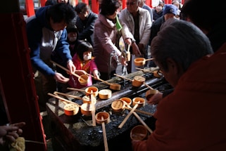 A group of people, including adults and children, are gathered around a traditional water purification fountain. They are holding wooden ladles used to scoop and pour water as part of a cleansing ritual. The atmosphere appears busy and communal, with everyone actively engaged in the purification process.