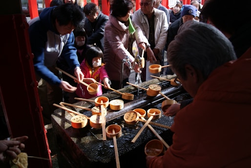 A group of people, including adults and children, are gathered around a traditional water purification fountain. They are holding wooden ladles used to scoop and pour water as part of a cleansing ritual. The atmosphere appears busy and communal, with everyone actively engaged in the purification process.