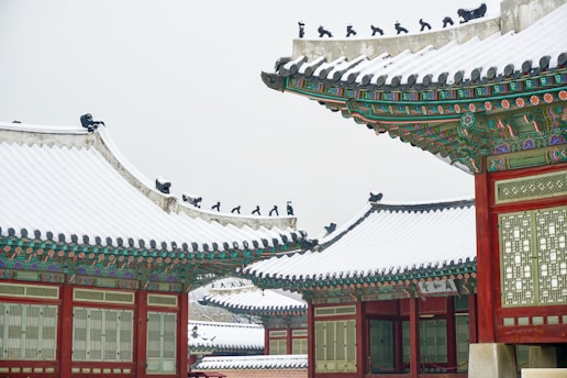 Ornate traditional Asian architecture featuring intricately designed roofs with vibrant green, red, and gold accents. Snow blankets the rooftops, adding a serene winter atmosphere. Decorative figures line the ridges of the roofs, enhancing the cultural aesthetics.