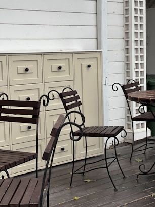 A set of wrought iron chairs with wooden slats is positioned on a wooden deck. Beige cabinets with multiple drawers add a vintage touch to the outdoor setting. The area includes white panel walls and a wooden trellis, enhancing the cozy and rustic atmosphere.