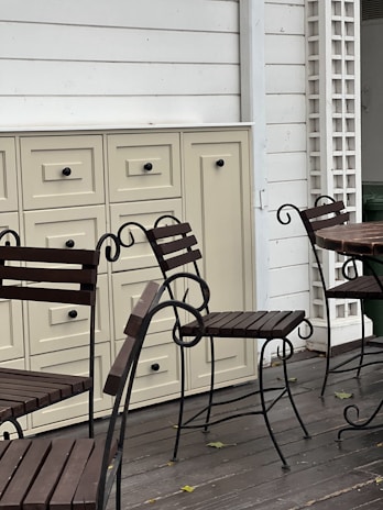 A set of wrought iron chairs with wooden slats is positioned on a wooden deck. Beige cabinets with multiple drawers add a vintage touch to the outdoor setting. The area includes white panel walls and a wooden trellis, enhancing the cozy and rustic atmosphere.