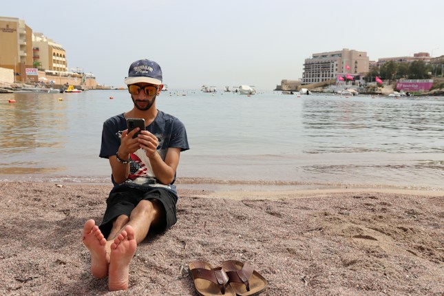 A man is sitting barefoot on a sandy beach, holding a smartphone. He's wearing sunglasses, a cap, and a casual t-shirt. In the background, there are several buildings, and boats are floating on calm water.