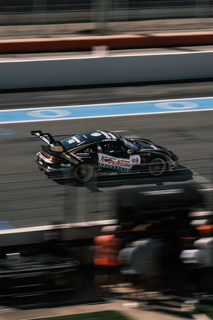 A sleek race car is speeding down a professional racing track. The car is black with prominent sponsor logos, including the number 98, and features a large rear spoiler. The background shows blurred motion lines indicating high speed, and part of the pit lane with indistinct figures wearing safety vests.