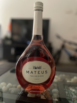 A bottle of Mateus Rosé wine with a uniquely shaped, elegant glass bottle. The label features the words 'Mateus The Original Rosé Portugal' along with an illustration of an estate. The bottle is on a glass table with decorative stones underneath, and the background is slightly blurred.