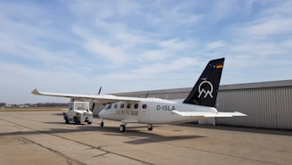 A small white aircraft with 'ALPEN AIR' written on its side is parked on a tarmac. The tail of the plane is black with a logo and a German flag. It is connected to an aircraft tug vehicle, which is positioned in front of it. The background includes a large hangar and a cloudy sky.