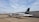 A small white aircraft with 'ALPEN AIR' written on its side is parked on a tarmac. The tail of the plane is black with a logo and a German flag. It is connected to an aircraft tug vehicle, which is positioned in front of it. The background includes a large hangar and a cloudy sky.