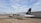 A small white aircraft with 'ALPEN AIR' written on its side is parked on a tarmac. The tail of the plane is black with a logo and a German flag. It is connected to an aircraft tug vehicle, which is positioned in front of it. The background includes a large hangar and a cloudy sky.