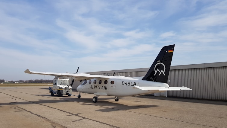A small white aircraft with 'ALPEN AIR' written on its side is parked on a tarmac. The tail of the plane is black with a logo and a German flag. It is connected to an aircraft tug vehicle, which is positioned in front of it. The background includes a large hangar and a cloudy sky.