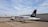 A small white aircraft with 'ALPEN AIR' written on its side is parked on a tarmac. The tail of the plane is black with a logo and a German flag. It is connected to an aircraft tug vehicle, which is positioned in front of it. The background includes a large hangar and a cloudy sky.