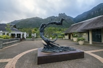A stylized sculpture of a leaping animal, possibly an antelope, displayed on a plinth in an open courtyard. The setting includes thatched-roof buildings and large potted plants, with a backdrop of lush green mountains under a cloudy sky.