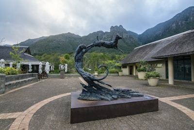 A stylized sculpture of a leaping animal, possibly an antelope, displayed on a plinth in an open courtyard. The setting includes thatched-roof buildings and large potted plants, with a backdrop of lush green mountains under a cloudy sky.