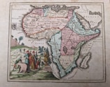 A historical map of the African continent with colorful borders that separate various regions. Detailed labels of regions, cities, and geographical features are visible. Near the bottom-left corner, illustrations of people wearing historical attire alongside an elephant and other fauna are depicted. There is an artistic rendering of landscapes in the background including trees and mountains.
