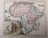 A historical map of the African continent with colorful borders that separate various regions. Detailed labels of regions, cities, and geographical features are visible. Near the bottom-left corner, illustrations of people wearing historical attire alongside an elephant and other fauna are depicted. There is an artistic rendering of landscapes in the background including trees and mountains.