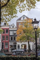 A street scene with traditional European row houses featuring colorful facades, large windows, and a decorative gable roof. The foreground includes a tree with autumn foliage and a parked bicycle. A car is visible on the street alongside a café with a sign reading 'GRAB LIFE BY THE BEANS'. A streetlamp and some tents or covered stalls appear on the sidewalk.