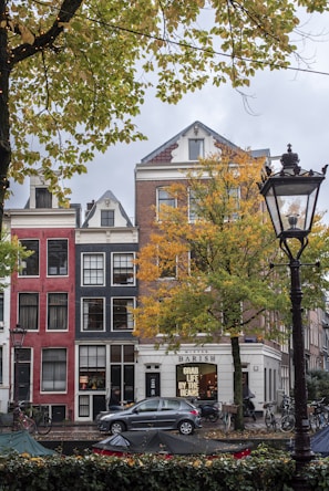 A street scene with traditional European row houses featuring colorful facades, large windows, and a decorative gable roof. The foreground includes a tree with autumn foliage and a parked bicycle. A car is visible on the street alongside a café with a sign reading 'GRAB LIFE BY THE BEANS'. A streetlamp and some tents or covered stalls appear on the sidewalk.