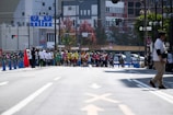 A busy city street is the setting for a sporting event. Groups of athletes, some in wheelchairs, are positioned at the starting line, dressed in colorful uniforms. Spectators and officials in casual attire can be seen along the sidelines. The street is lined with traffic cones, and signs with Japanese writing are visible in the background, hinting at an urban location. Trees and buildings fill the backdrop, with a variety of traffic and directional signs adding to the urban environment.