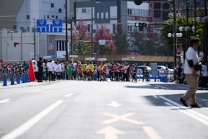 A busy city street is the setting for a sporting event. Groups of athletes, some in wheelchairs, are positioned at the starting line, dressed in colorful uniforms. Spectators and officials in casual attire can be seen along the sidelines. The street is lined with traffic cones, and signs with Japanese writing are visible in the background, hinting at an urban location. Trees and buildings fill the backdrop, with a variety of traffic and directional signs adding to the urban environment.