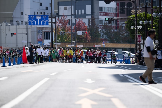A busy city street is the setting for a sporting event. Groups of athletes, some in wheelchairs, are positioned at the starting line, dressed in colorful uniforms. Spectators and officials in casual attire can be seen along the sidelines. The street is lined with traffic cones, and signs with Japanese writing are visible in the background, hinting at an urban location. Trees and buildings fill the backdrop, with a variety of traffic and directional signs adding to the urban environment.