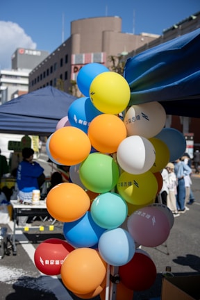 A cluster of colorful balloons is tied to a metal stand at an outdoor event. The balloons come in shades of yellow, blue, orange, green, red, and white, with some featuring printed text. People are visible in the background, along with tents and urban buildings under a clear blue sky.