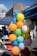 A cluster of colorful balloons is tied to a metal stand at an outdoor event. The balloons come in shades of yellow, blue, orange, green, red, and white, with some featuring printed text. People are visible in the background, along with tents and urban buildings under a clear blue sky.
