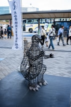 A wire sculpture resembling a creature is displayed on a platform in a busy outdoor area. People walk by in the background, while a bus is parked nearby. A tall banner with text stands behind the sculpture.