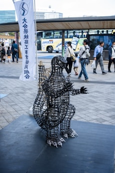 A wire sculpture resembling a creature is displayed on a platform in a busy outdoor area. People walk by in the background, while a bus is parked nearby. A tall banner with text stands behind the sculpture.