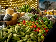 A vibrant display of various fresh vegetables and grains is laid out on a market stall. There are bright green okra, an assortment of bell peppers, and plastic bags filled with grains like corn and beans. In the background, bananas sit in a bunch, while larger grains and fruits are visible, contributing to a lively market atmosphere.