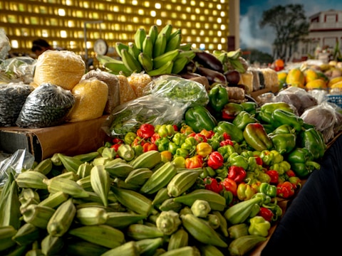 A vibrant display of various fresh vegetables and grains is laid out on a market stall. There are bright green okra, an assortment of bell peppers, and plastic bags filled with grains like corn and beans. In the background, bananas sit in a bunch, while larger grains and fruits are visible, contributing to a lively market atmosphere.