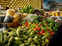 A vibrant display of various fresh vegetables and grains is laid out on a market stall. There are bright green okra, an assortment of bell peppers, and plastic bags filled with grains like corn and beans. In the background, bananas sit in a bunch, while larger grains and fruits are visible, contributing to a lively market atmosphere.
