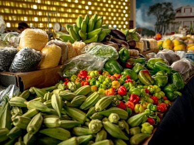 A vibrant display of various fresh vegetables and grains is laid out on a market stall. There are bright green okra, an assortment of bell peppers, and plastic bags filled with grains like corn and beans. In the background, bananas sit in a bunch, while larger grains and fruits are visible, contributing to a lively market atmosphere.
